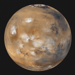 Figure 5.3. This is a global view of Mars. Many different geologic features can be distinguished on the planet’s surface of mostly red-orange color. Huge volcanoes up to 24 km in height above the mean surface level are seen in the elevated Tharsis region at the left. They are partially obscured by the clouds in the very rarefied atmosphere (white patches). The Valley Marineris, some hundreds of kilometers wide and 8 km deep, extends from just South of the equator for more than 3000 km. Cratered terrain is seen at higher southern latitudes. At the top of the image is the North polar cap, composed of water ice and overlying frozen carbon dioxide (“dry ice” deposits) formed during the Martian winter. (Courtesy of NASA)